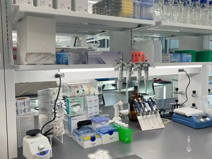 A laboratory bench with a bound laboratory notebook open beside a pipette rack, reagent bottles and a vortex mixer