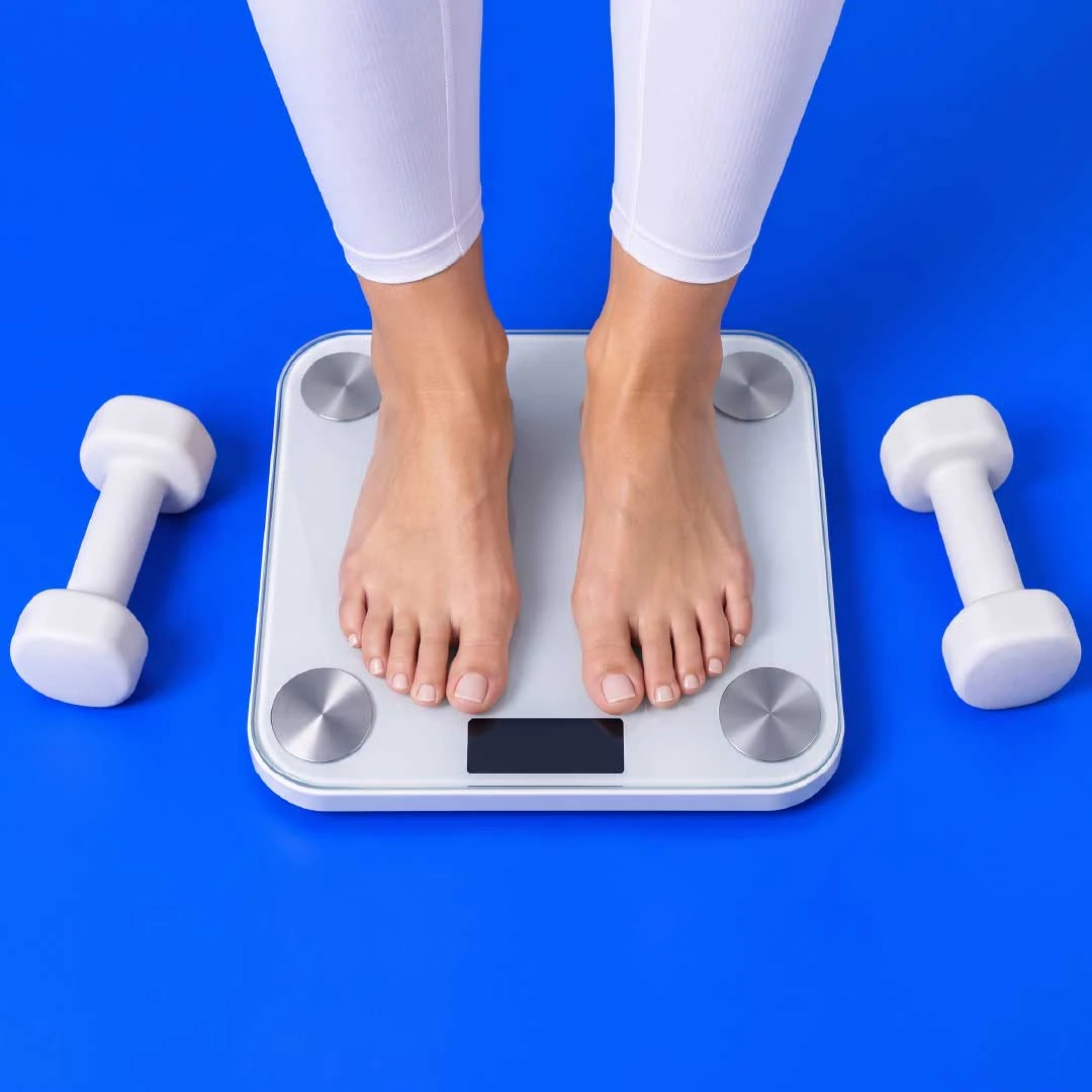 Bare feet on a white bathroom scale between two white dumbbells on a blue ground